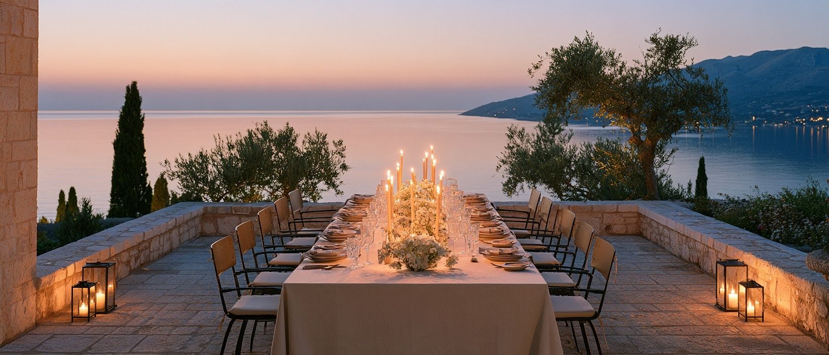 A long candlelit dinner table on a stone terrace above the sea at dusk